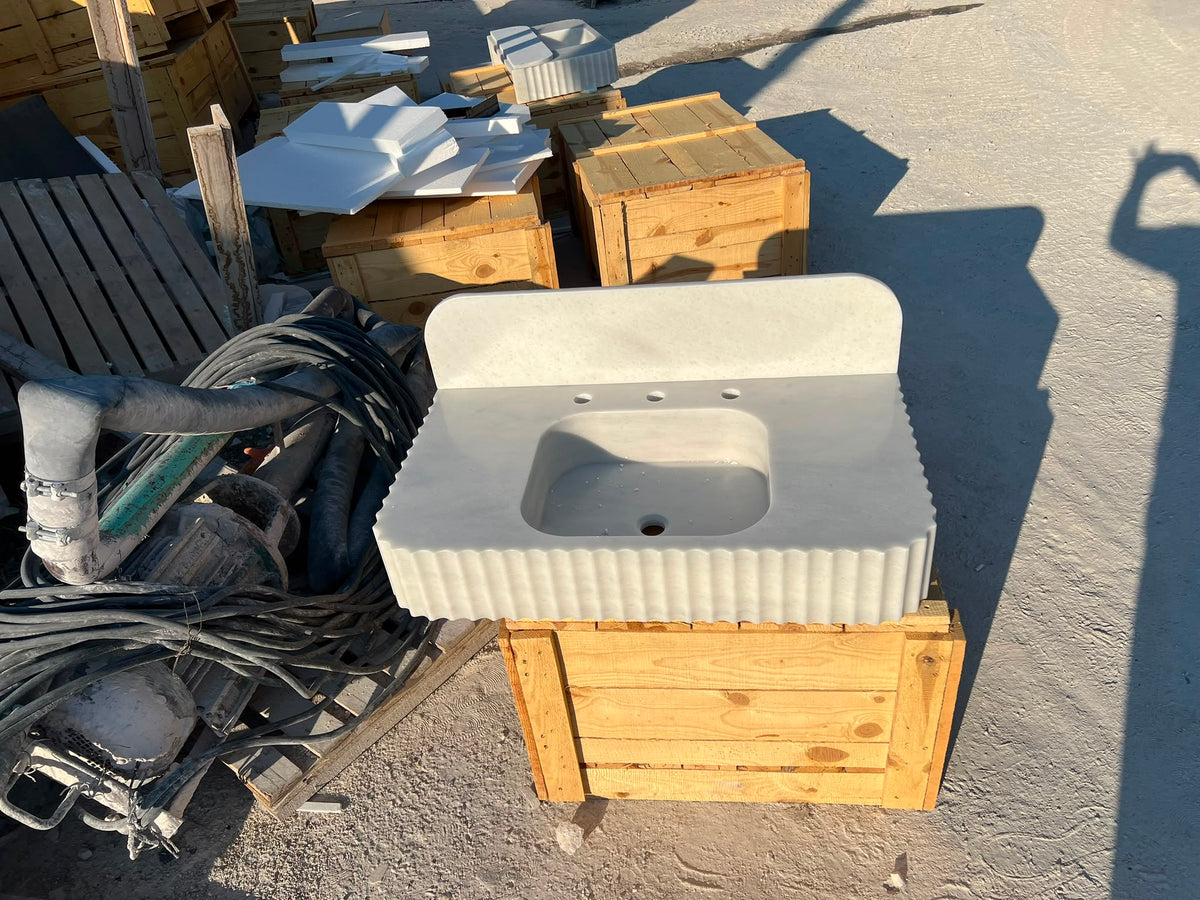 A white carrara marble bathroom vanity sink top with a fluted front and integrated basin, displayed outdoors on a wooden support crate in a workshop or storage area.